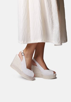 Nohy v bielych sandáloch na kline so zapínaním za pätou a pletenou podrážkou z espadril, v kombinácii s bielou textúrovanou midi sukňou.