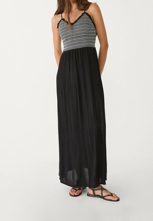 Woman wearing black maxi dress with white geometric pattern on bodice and black strappy flat sandals on plain background.