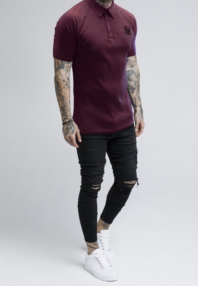 Homme portant un polo ajusté bordeaux, un jean skinny noir déchiré, des baskets blanches, avec des tatouages sur les bras jusqu'aux poignets.