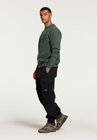 Felpa verde con logo rosso, pantaloni cargo neri con tasche laterali e sneaker New Balance verde oliva con dettagli rosa.