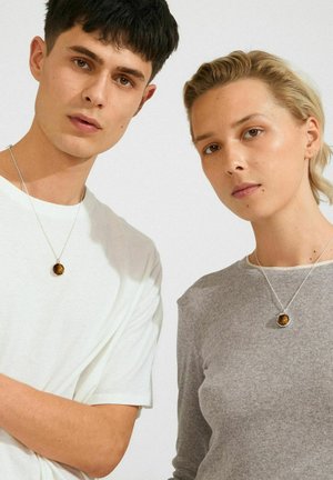 Two young adults wearing similar tiger's eye stone pendant necklaces, one in a white shirt and one in a gray long-sleeve top, neutral background.