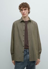 Oliwkowa zielona koszula z kołnierzykiem, długimi rękawami i zapięciem na guziki z przodu. Tkanina ma delikatną fakturę, w połączeniu z brązową koszulą pod spodem.