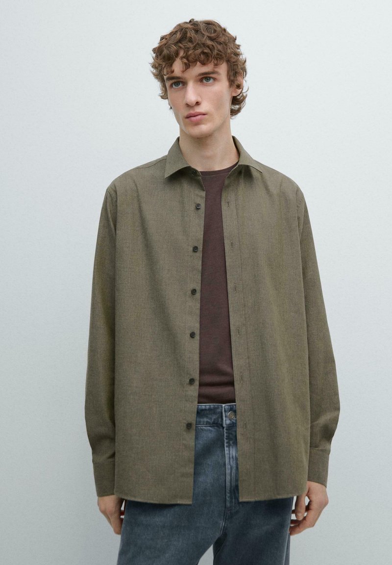 Oliwkowa zielona koszula z kołnierzykiem, długimi rękawami i zapięciem na guziki z przodu. Tkanina ma delikatną fakturę, w połączeniu z brązową koszulą pod spodem.