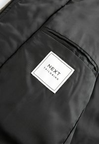 Tissu noir à texture lisse avec une étiquette blanche. L'étiquette porte l'inscription "NEXT TAILORING" en texte noir gras, avec une petite poche à proximité.