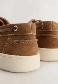 Braune Wildlederschuhe mit einem niedrigen Schnitt, ausgestattet mit weißen Nähten und Gummisohlen. Die Absätze sind rund und haben Ösen für die Schnürsenkel.