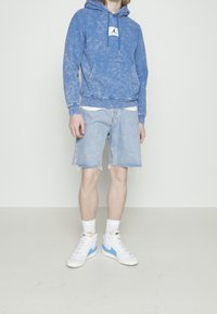 Sudadera azul con efecto tie-dye y cordones, pantalones cortos de mezclilla con el dobladillo deshilachado y zapatillas blancas con acentos azules. Camiseta blanca lisa visible.