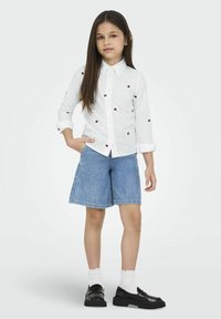 Camisa blanca de botones con bordado de un corazón rojo, combinada con pantalones cortos de mezclilla azul. El calzado son zapatos negros, complementados con calcetines blancos hasta el tobillo.