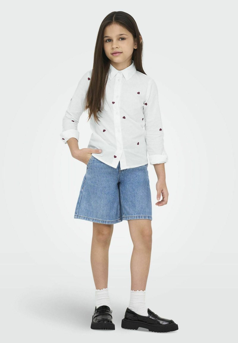Camisa blanca de botones con bordado de un corazón rojo, combinada con pantalones cortos de mezclilla azul. El calzado son zapatos negros, complementados con calcetines blancos hasta el tobillo.