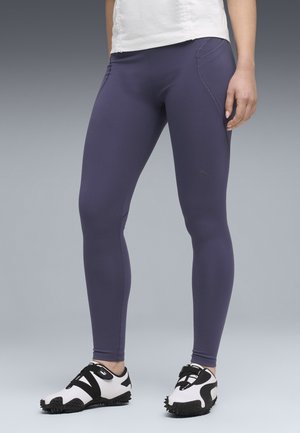 Piernas con leggings deportivos de color púrpura oscuro y zapatillas deportivas blancas Puma con suelas y correas negras, de pie sobre una superficie gris claro.