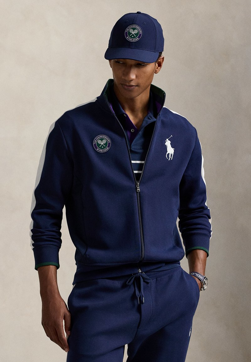 Tmavomodrá bunda na zips s bielymi bočnými panelmi, vyšívané logo Wimbledon na hrudi, v kombinácii s pruhovaným polokošeľou a zodpovedajúcimi športovými nohavicami. Tmavomodrá čiapka.