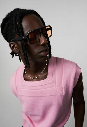 Lunettes de soleil noires surdimensionnées avec des verres ambre, débardeur en tricot rose et collier en perles argentées, sur un fond neutre.
