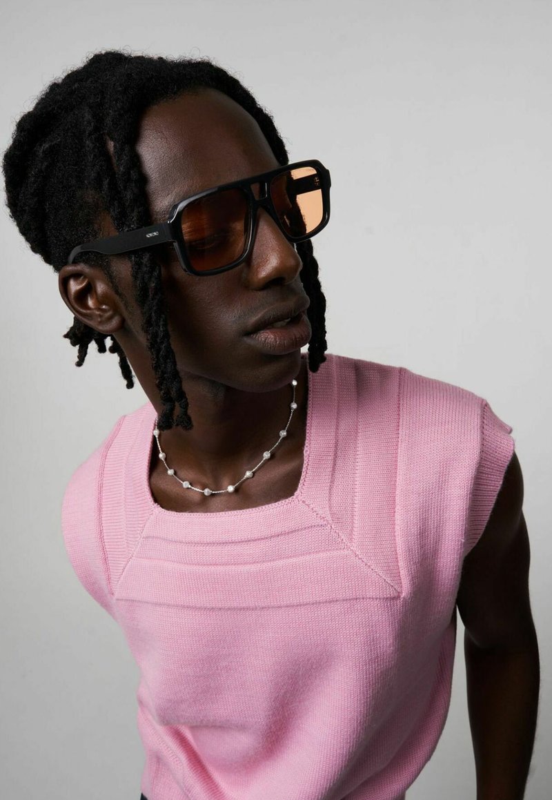 Occhiali da sole neri oversize con lenti ambra, top smanicato lavorato a maglia rosa e collana di perline argentate, su uno sfondo neutro.