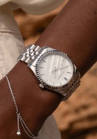 Silberner Edelstahl-Armbanduhr mit Zifferblatt aus weißem Perlmutt, dünnen Stundenmarkierungen und einer strukturierten Lünette, getragen an einem braunen Handgelenk.