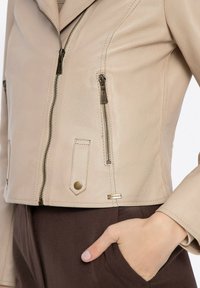 Beige leren jasje met een getailleerd ontwerp, voorzien van twee met rits afgesloten zakken en een decoratieve drukknoop aan de voorkant. Geweven textuur met zichtbare stiksels.