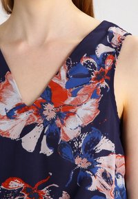 Blouse florale avec un décolleté en V profond, fabriquée en tissu léger. Le design comprend de grandes fleurs blanches, rouges et bleues sur un fond sombre.