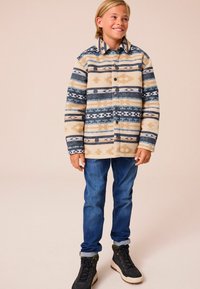 Beżowa flanelowa kurtka z niebieskimi i brązowymi geometrycznymi wzorami, z przodu zapinana na guziki oraz kołnierzem, w zestawieniu z niebieskimi dżinsami i czarnymi butami.