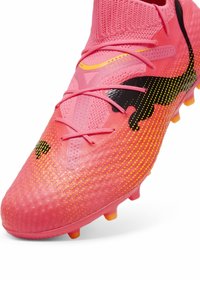 Botín de fútbol de color rosa brillante con superficie texturizada, acentos negros y amarillos, y un ajuste ceñido similar a un calcetín. Presenta un sistema de cordones tradicional.