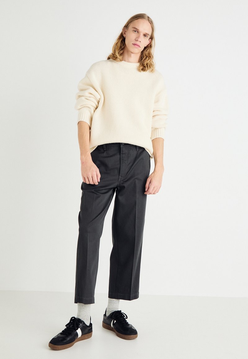 Suéter oversize crema, pantalones de vestir negros y zapatillas negras con acentos blancos. Calcetines gris claro visibles por encima de los zapatos.