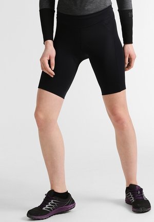 Personne vêtue d'un short de cyclisme noir, d'un haut gris à manches longues et de chaussures de course noir-violet, debout, jambes écartées.
