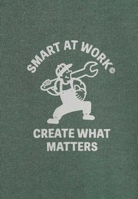 Tessuto verde con design grafico bianco che raffigura un personaggio dei cartoni animati che tiene una chiave inglese. Il testo recita "SMART AT WORK" e "CREATE WHAT MATTERS."
