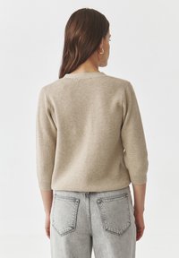 Beige pruhovaný svetr s tříčtvrtečními rukávy, pohled zezadu. Měkká textura; kulatý výstřih. V kombinaci s vysokými světle šedými džíny.