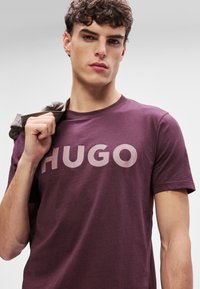 Bordo t-särk, millel on suur heledoosa "HUGO" logo. Kangas näeb välja pehme ja sellel on ümar kaelus. Õlavarre peal on heledat halli värvi jakk.