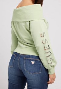 Femme portant un haut vert clair à épaules dénudées avec le texte "GUESS" sur la manche et un jean bleu avec un logo triangle Guess sur la poche arrière.