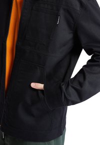 Černá džínsová bunda se zipem vpředu, dvěma náprsními kapsami a jednou boční kapsou. Obsahuje oranžovou podšívku a štítek značky na boku.