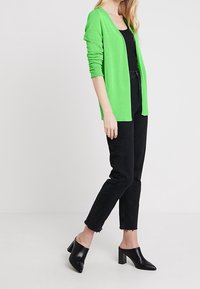 Bright green long-sleeve cardigan over a black tank top, paired with high-waisted black jeans and black open-toe heels.