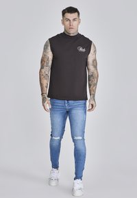 Black sleeveless top with high collar, paired with light blue distressed skinny jeans and white sneakers. Model has tattoos and short hair.