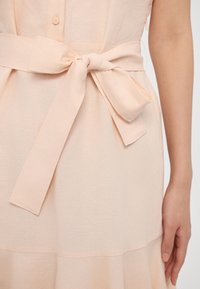 Robe pêche clair avec une finition texturée, dotée d'une ceinture nouée à la taille et de détails en boutons sur le devant. Tissu doux avec une jupe fluide.