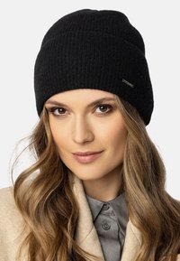 Gorro de punto negro con textura acanalada, que presenta un pequeño detalle de etiqueta metálica. Lo lleva puesto una persona con cabello largo y una chaqueta de color claro.
