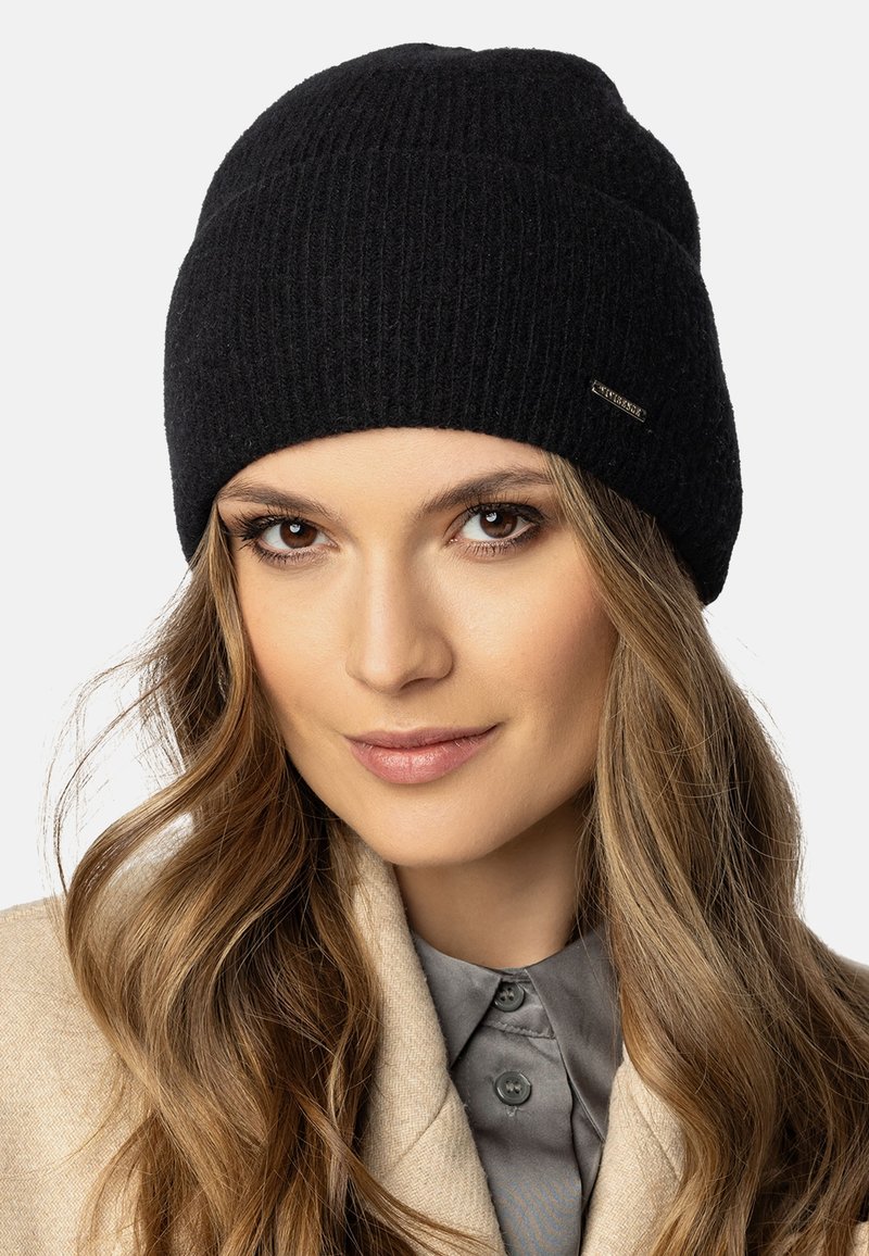 Gorro de punto negro con textura acanalada, que presenta un pequeño detalle de etiqueta metálica. Lo lleva puesto una persona con cabello largo y una chaqueta de color claro.