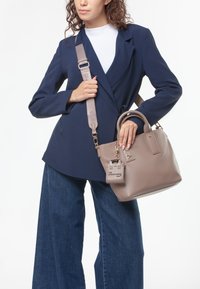 Tan handtas met dubbele handgrepen en een afneembare schouderriem, met een label waarop staat "IK BEN EEN VERANTWOORD BAG", tegen een marineblauwe blazer.