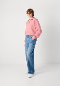 Pullover corto rosa chiaro con colletto, abbinato a jeans larghi azzurri. Calzature sono sneakers bianche. Accentuato con occhiali da sole rotondi.