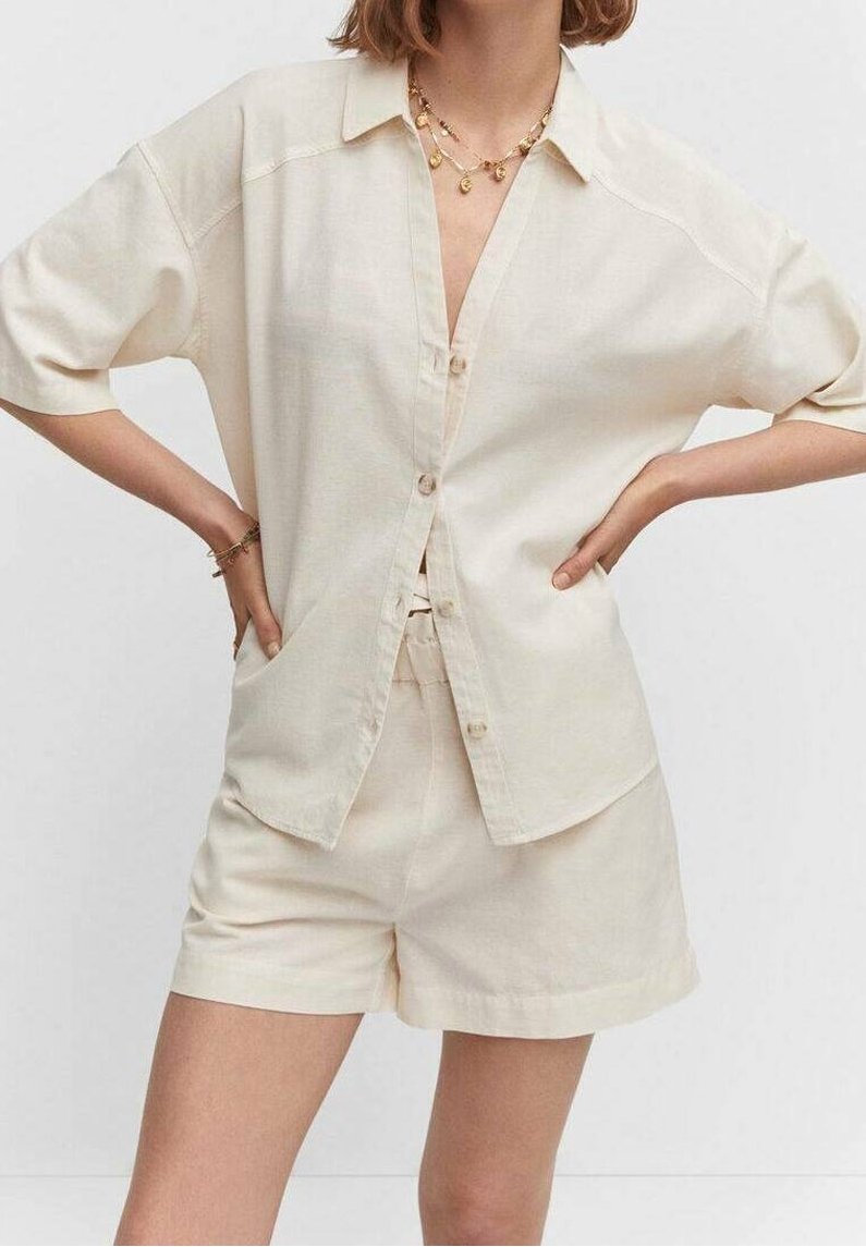 Femme portant une chemise crème à manches courtes boutonnée et un short assorti, accessoirisée avec des colliers et bracelets en or superposés, mains sur les hanches.