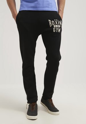 Mann trägt schwarze Jogginghose mit dem Schriftzug „Boxing Gym“ am linken Oberschenkel, blaues Shirt und graue Sneakers mit braunen Schnürsenkeln und steht mit den Händen in den Taschen.