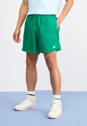 Shorts in nylon verde con cintura regolabile e tasche laterali, presentano un logo Nike bianco sulla gamba sinistra. Indossati con sneakers bianche.