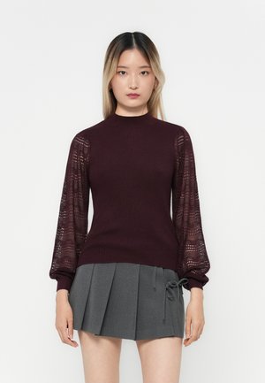 Bordeauxroter Strickpullover mit geripptem Körper und durchsichtigen, gemusterten Puffärmeln; kombiniert mit einem grauen, gefältelten Rock mit seitlichem Bindedetail.