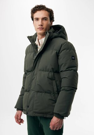 Giovane uomo con capelli ricci che indossa una giacca imbottita verde scuro con cappuccio, in piedi davanti a uno sfondo chiaro uniforme.