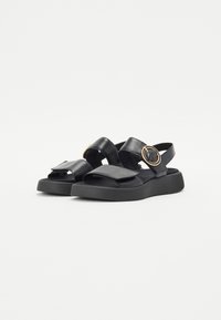 Black leather sandals with dual adjustable straps, a flat sole, and a circular metallic accent on the side. Textured footbed for comfort.