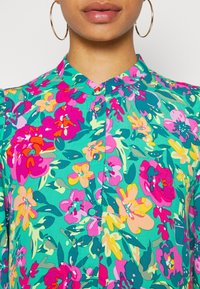 Camicia floreale multicolore con sfondo verde, fiori rosa e gialli, colletto e chiusura frontale con bottoni.