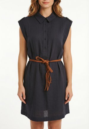 Mujer con vestido negro sin mangas abotonado con cuello y cinturón trenzado marrón atado a la cintura, parada contra un fondo blanco.