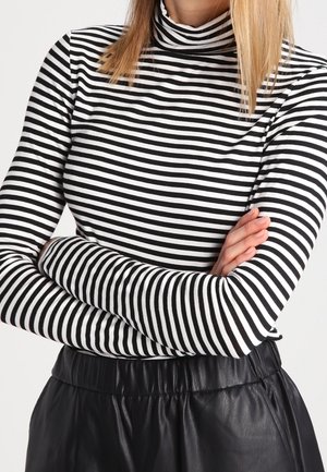 Vrouw met gekruiste armen, gekleed in een zwart-witte gestreepte coltrui en zwarte leren broek, tegen een effen achtergrond.