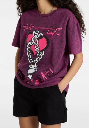 Person trägt ein lila T-Shirt mit einem Herz in Ketten-Grafik und schwarze Shorts, steht mit den Händen in den Taschen.