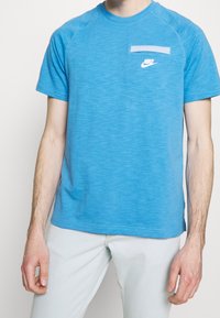 Camiseta de algodón azul con manga corta, cuello redondo y un bolsillo en el pecho. Presenta el logo de Nike en blanco y una textura en contraste en el cuello y las mangas.