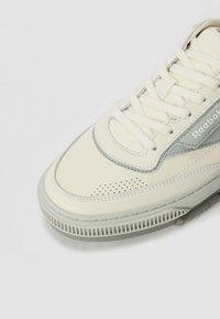 Cream-colored sneaker with a smooth leather upper, grey accents, perforated details, flat laces, and textured rubber sole.
