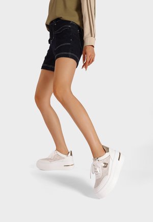 Sneakers bianchi con tomaia in rete e dettagli dorati. Suola spessa bianca, punta rotonda e stringhe. Indossati con pantaloni corti di jeans scuri e una maglietta verde.