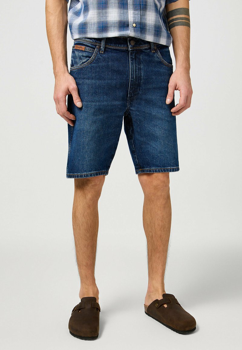 Jeans kratke hlače temno modre barve, ki imajo pet žepov in raztrgane robove. Nosi se jih z rjavimi lesnimi obuvali, v kombinaciji s karirasto srajco.