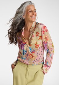 Smilende kvinde med langt gråt hår iført en farverig blomsterbluse og lysegrønne bukser, hænderne i lommerne, foran en hvid baggrund.